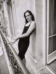 Model leaning against a stone building facade on a narrow balcony with ornate iron railing in a black and white editorial photograph.