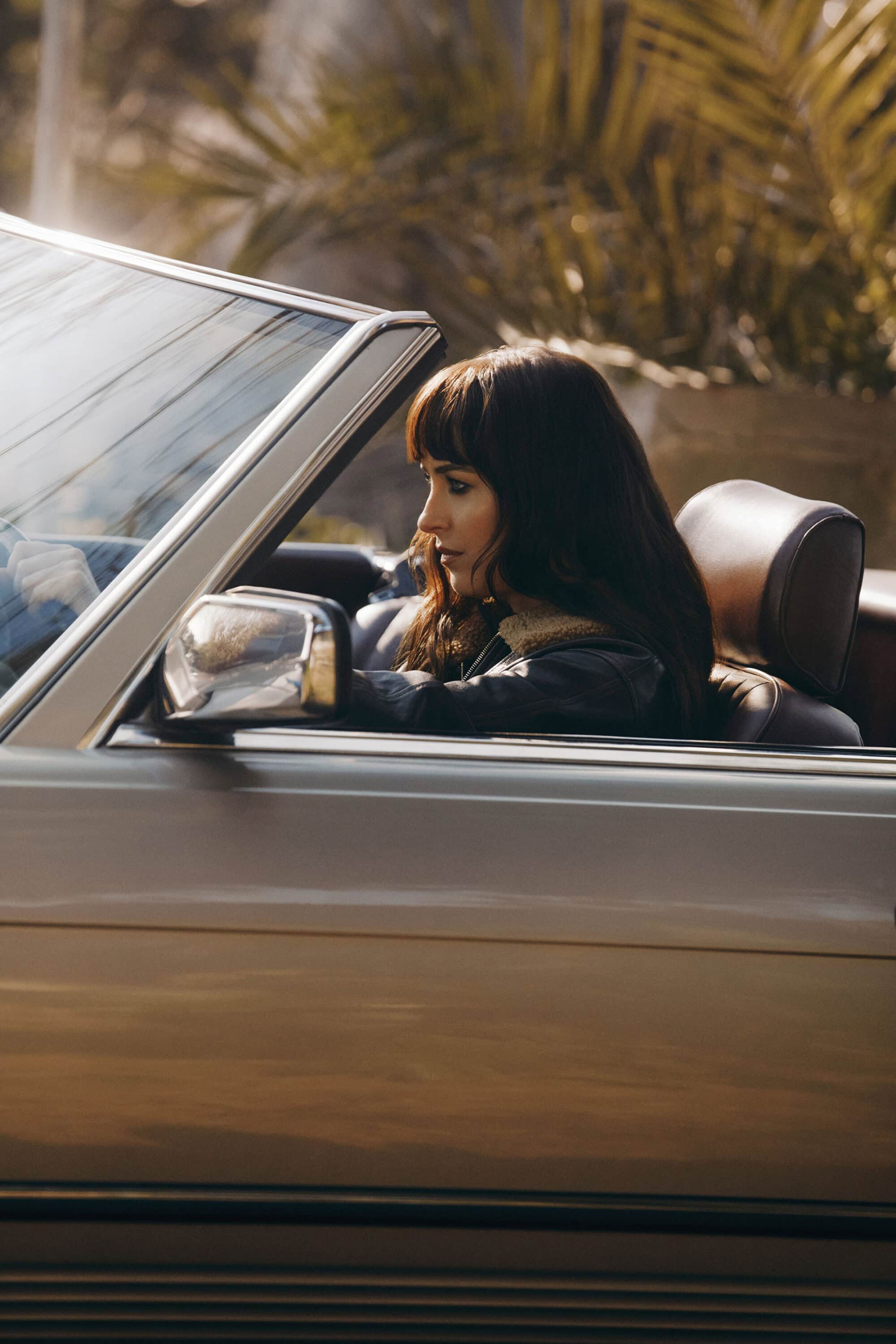 Dakota Johnson looking forward from the passenger seat of a convertible car outdoors in warm sunlight.