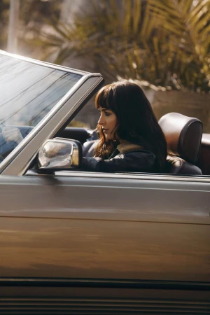 Dakota Johnson looking forward from the passenger seat of a convertible car outdoors in warm sunlight.