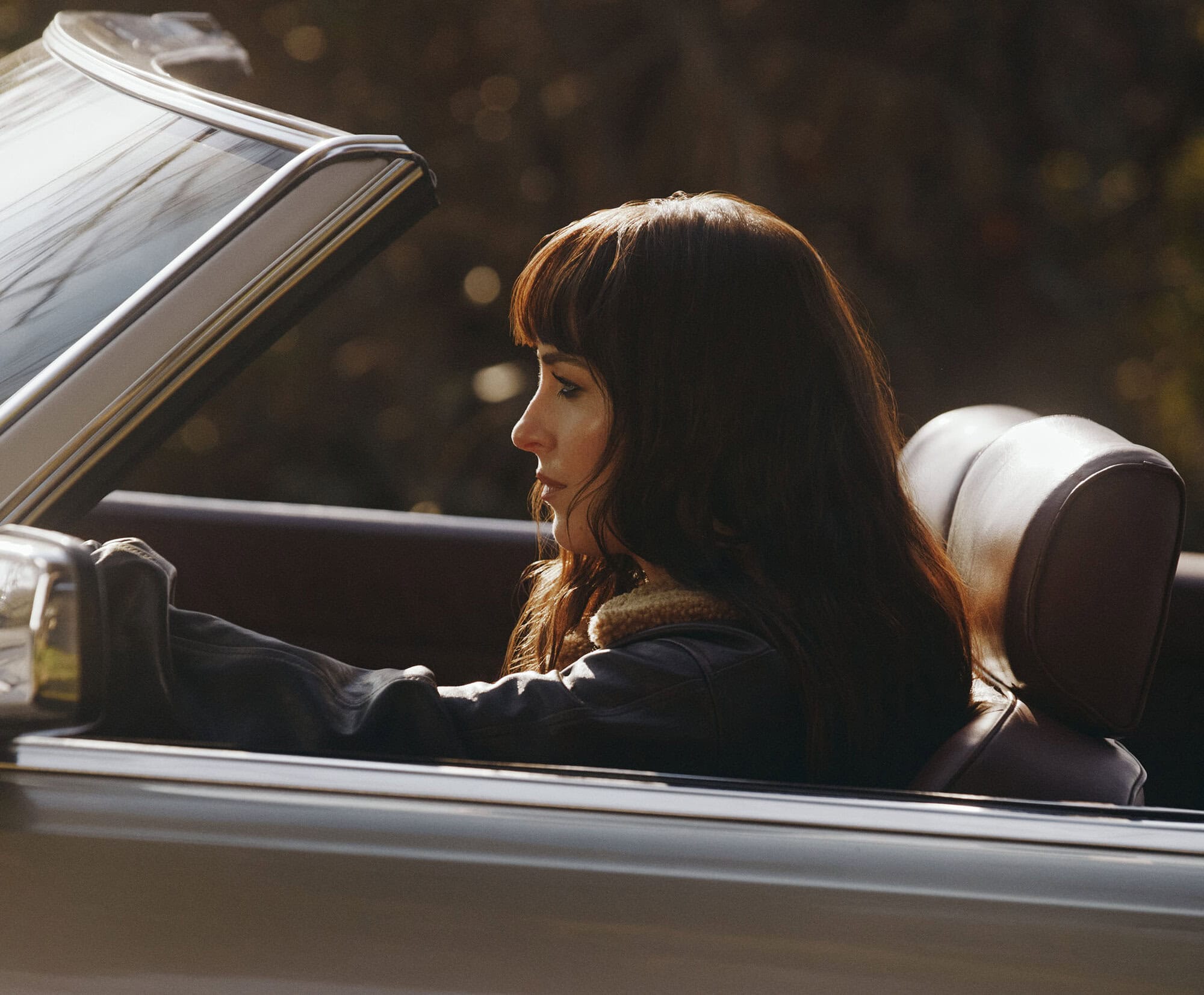 Dakota Johnson in profile driving a vintage convertible car wearing a dark jacket with a shearling collar.