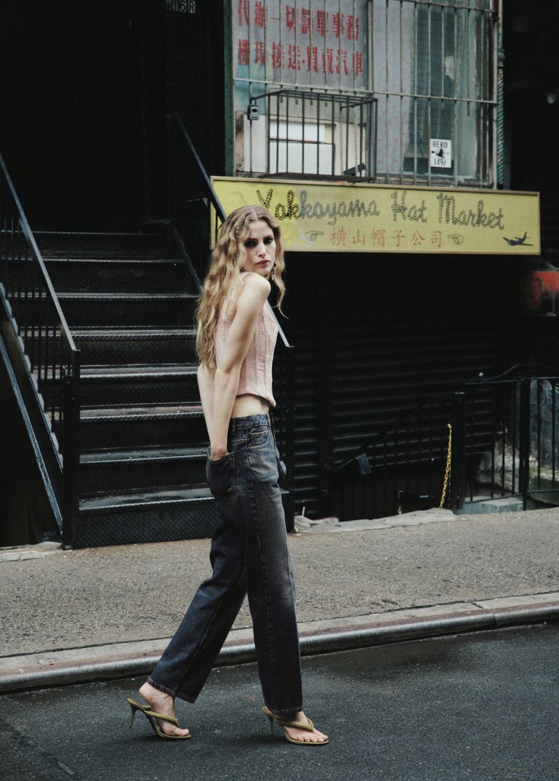 Model Felice Nova Noordhoff walks down a city street wearing a light pink top, dark wash denim jeans, and heeled sandals.