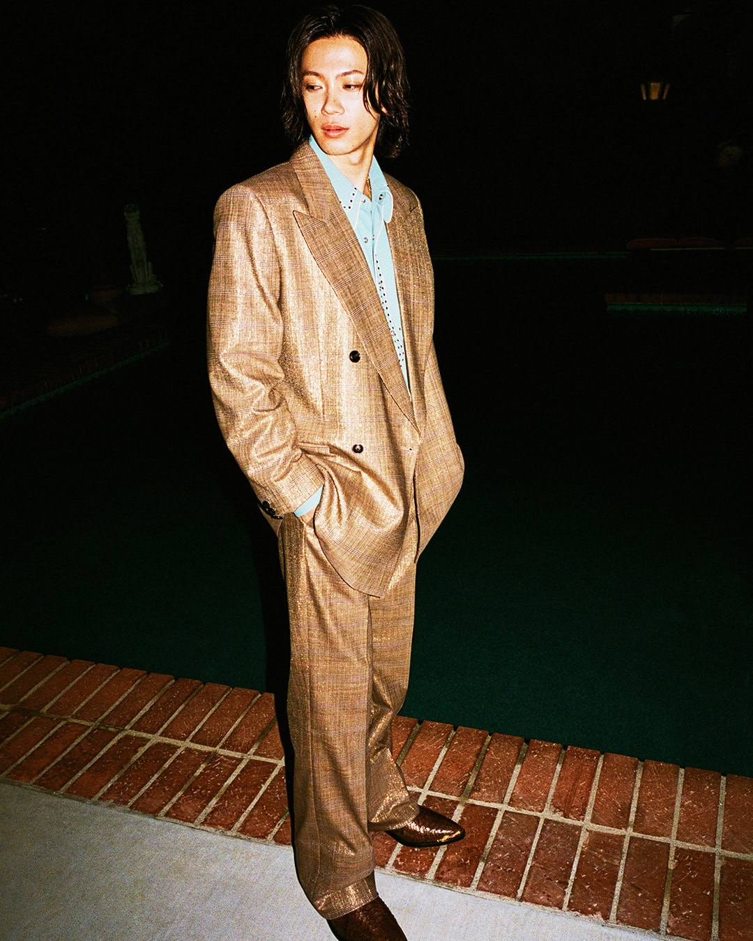 Yuta Jinguji standing poolside at night in an oversized brown tailored double-breasted suit and blue shirt.