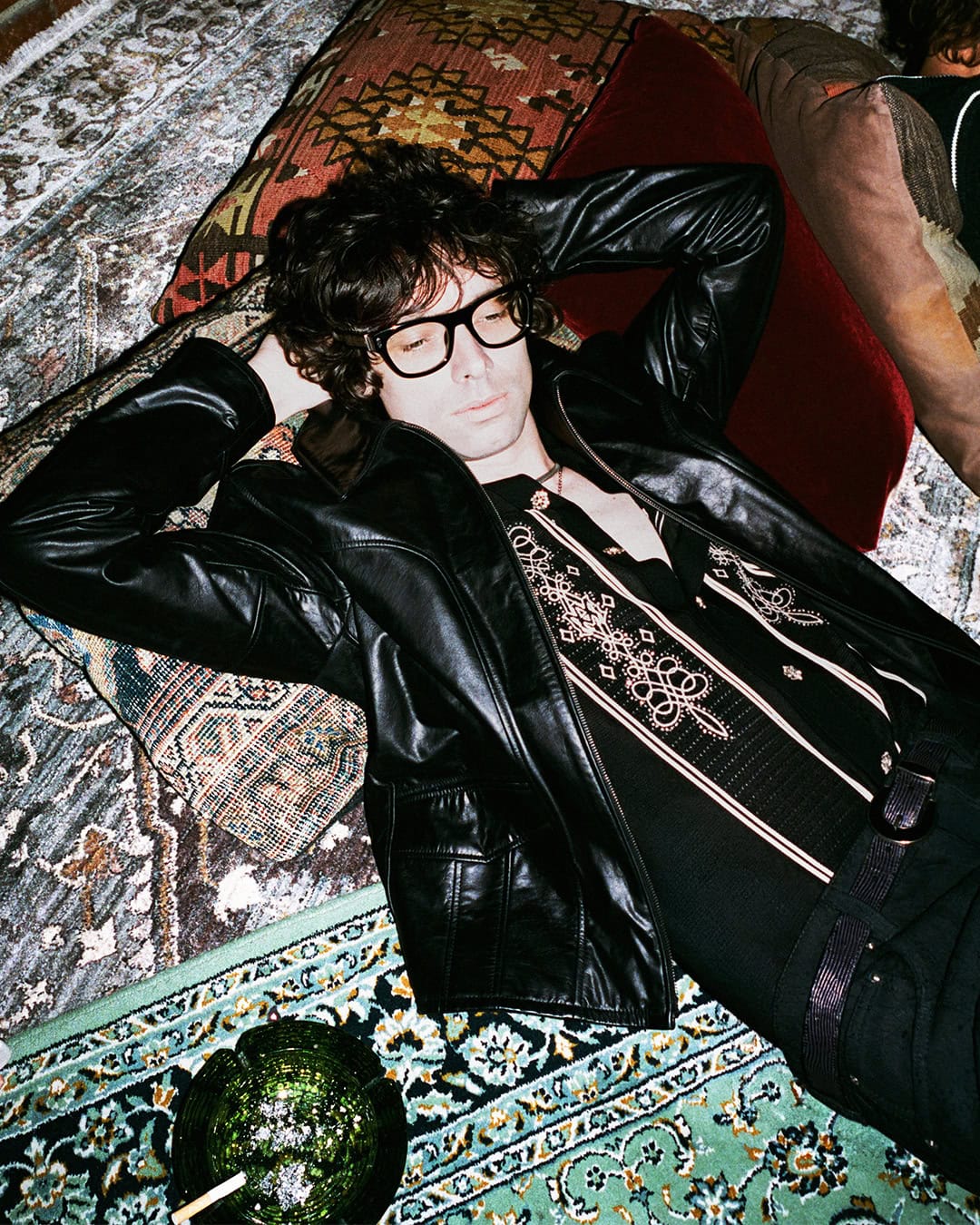 Man in black eyeglasses and a black leather jacket reclining back across layered patterned oriental rugs and cushions.