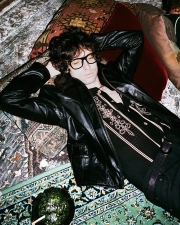 Man in black eyeglasses and a black leather jacket reclining back across layered patterned oriental rugs and cushions.