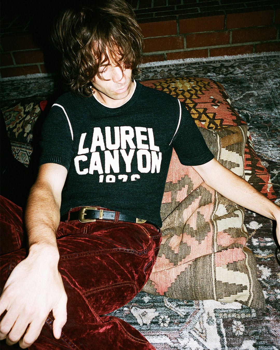 A man reclining on patterned floor cushions wearing a black Laurel Canyon knit shirt and burgundy velvet trousers.