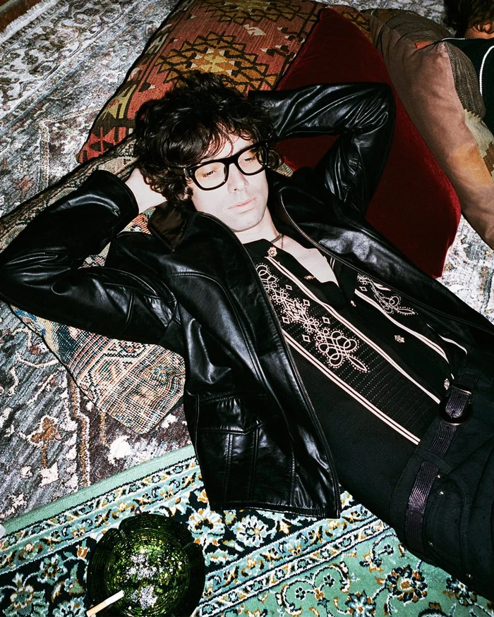 Man wearing black glasses, a leather jacket, and an embroidered shirt lies back on patterned carpets near a green glass ashtray.