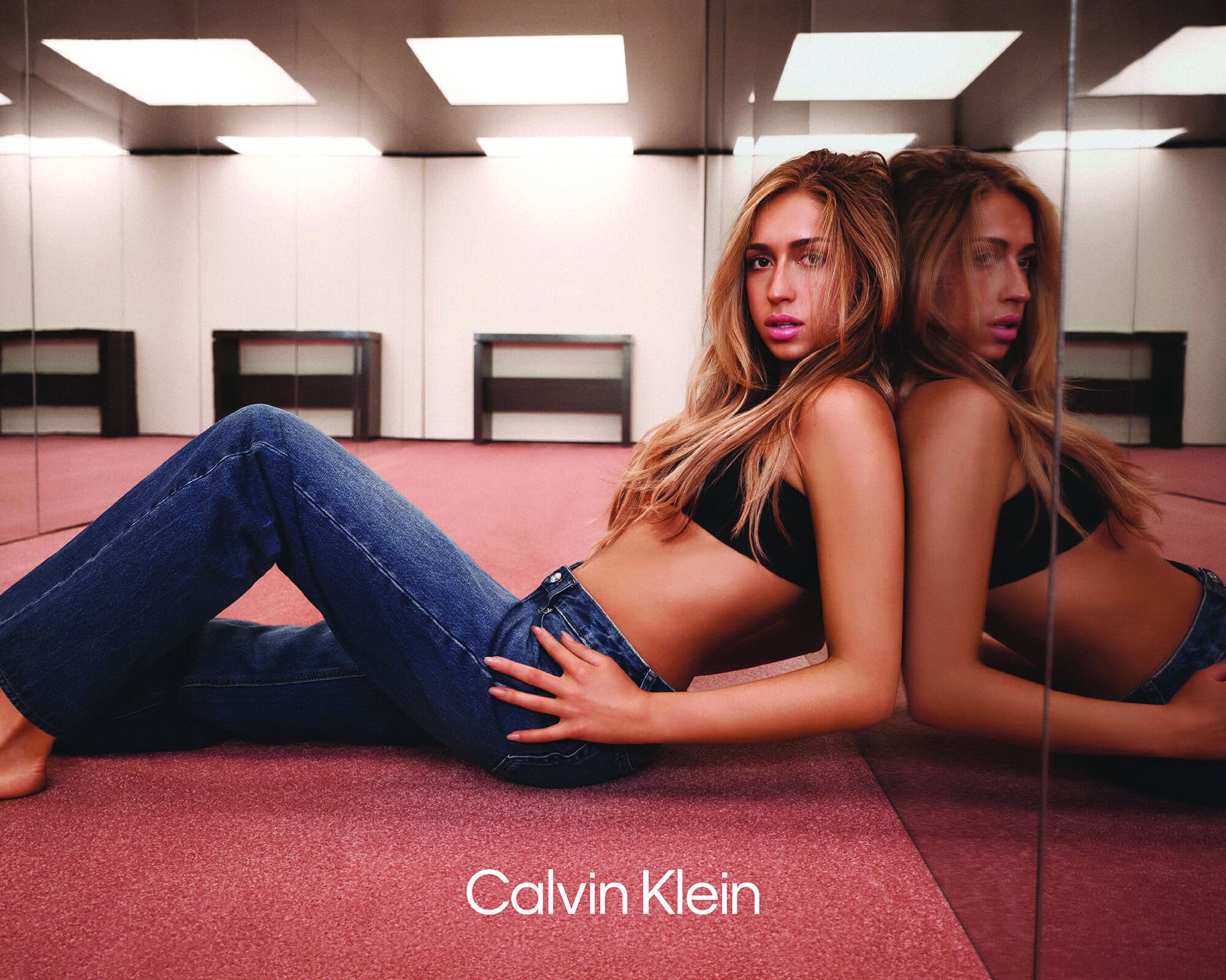 Tate McRae reclining on a pink carpet against a mirror wearing Calvin Klein blue jeans and a black bandeau top.