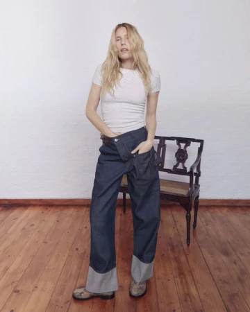 Dree Hemingway stands on a wood floor wearing a white t-shirt and wide-leg cuffed twisted denim jeans in front of an antique bench.