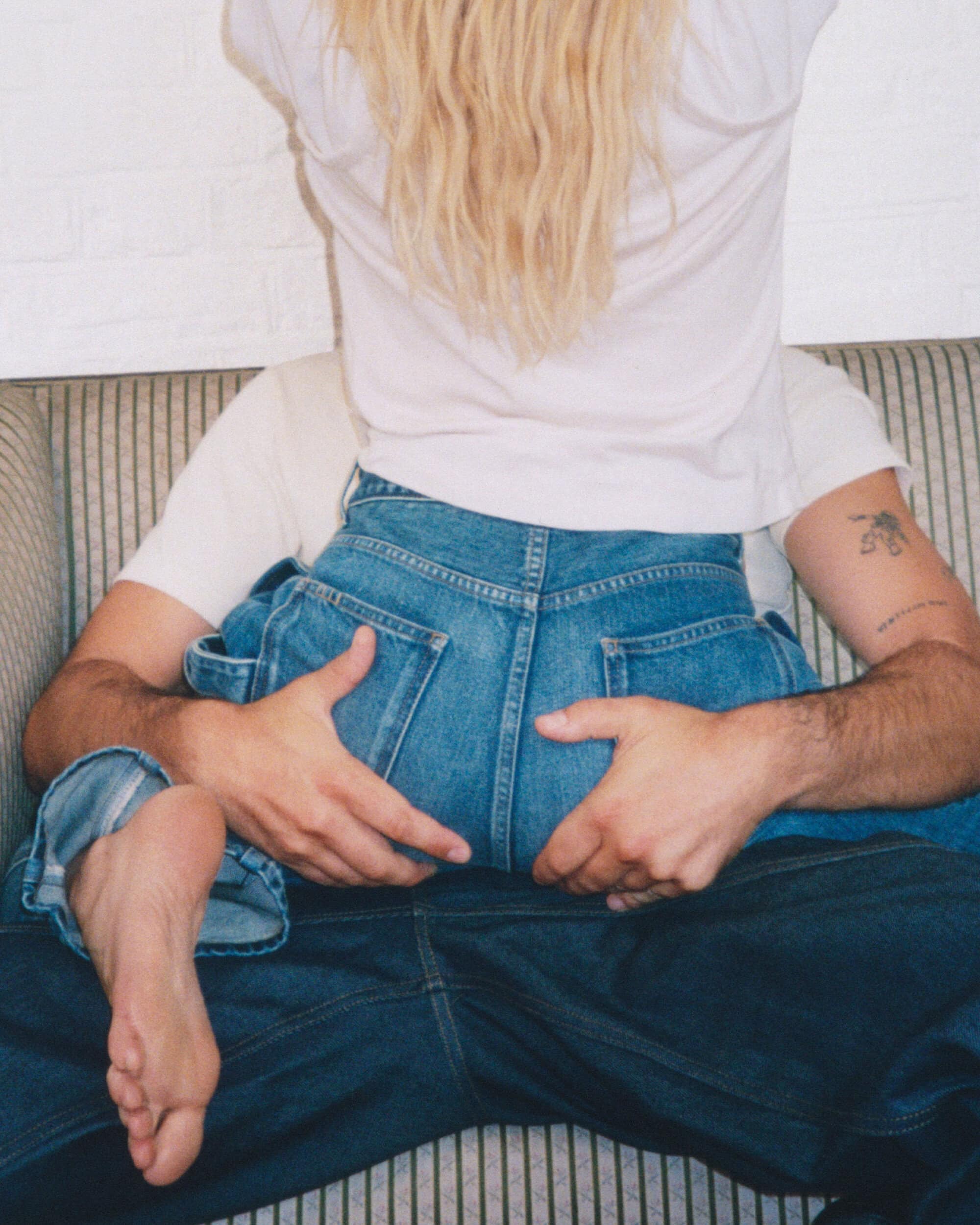 Dree Hemingway wearing blue denim jeans sitting on Nick Delli Santi's lap on a striped sofa for J.W. Anderson.