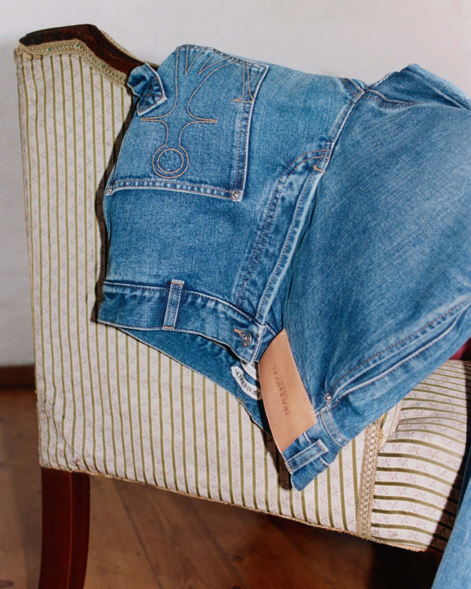 Blue denim jeans with embroidered anchor logo pocket draped over a vintage striped fabric armchair.