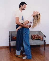 Nick Delli Santi holding Dree Hemingway leaning back in oversized denim jeans and cream t-shirts before a vintage sofa.