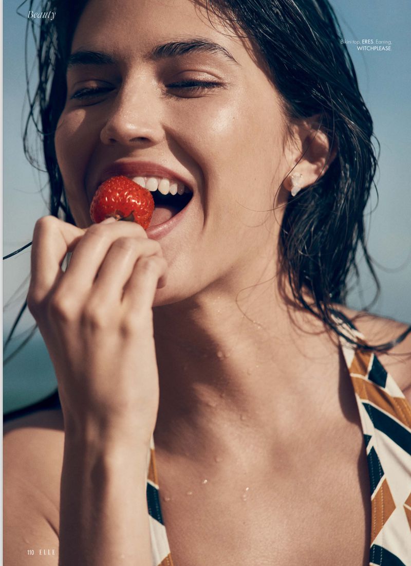 Smiling woman in swimwear eats a strawberry outdoors in soft light. Beauty shot with wet hair, showing skin texture and natural makeup.