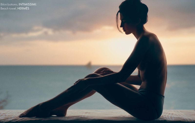 Silhouette of a young woman in swimwear sitting against a soft sunset beach backdrop, evoking peacefulness and a connection with nature.