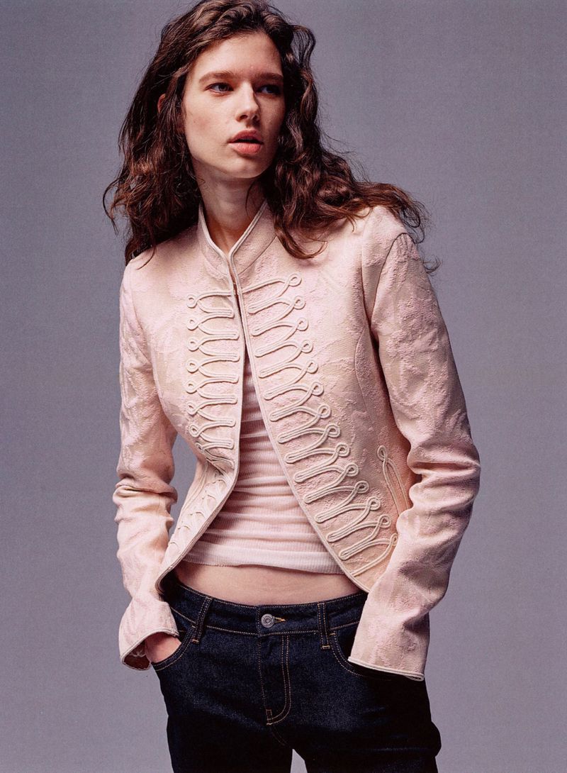 Model wearing a pale blush braided military style jacket and dark denim jeans against a solid grey studio background.