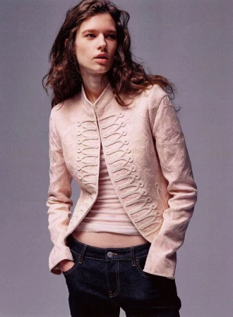 Model wearing a pale blush braided military style jacket and dark denim jeans against a solid grey studio background.
