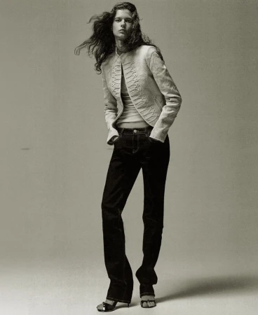 Model Felice Nova Noordhoff wearing a cream embroidered jacket and dark denim jeans standing against a grey studio backdrop.