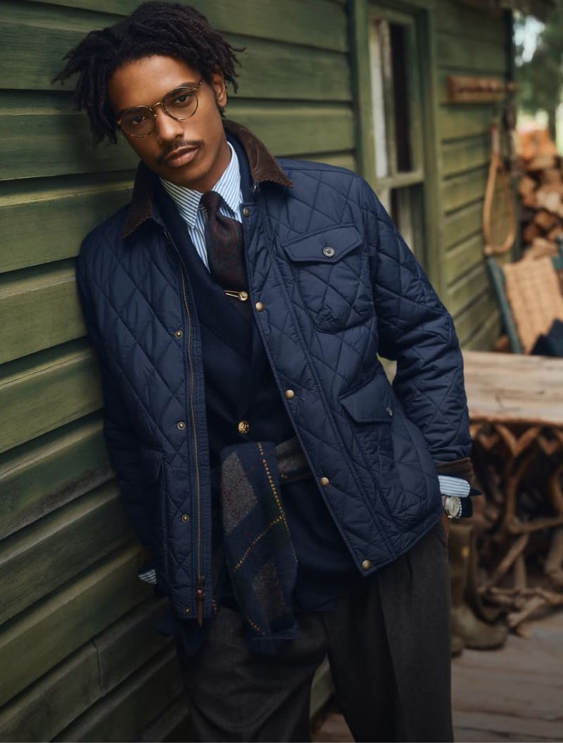Model Nile Winchester wearing glasses and a navy quilted jacket while leaning against a green wooden wall.