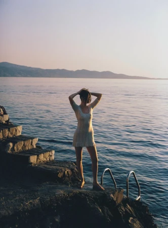 Model posing in a cream knit mini dress on a rocky sea ledge with calm water and distant mountains at sunset.