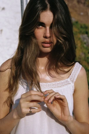 Zoi Aggeliki Mantzakanis holding a strand of hair wearing a white lace-trimmed camisole in natural direct sunlight.