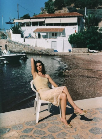 Model Zoi Aggeliki Mantzakanis sits on a white plastic chair by a coastal harbor wearing a pale yellow top and skirt.