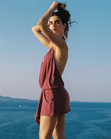 Model Zoi Aggeliki Mantzakanis posing in a mauve backless halter mini dress against a blue ocean backdrop.