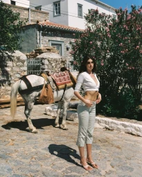 Model Zoi Aggeliki Mantzakanis wearing a white crop top and cropped trousers on a stone path next to a pack horse.