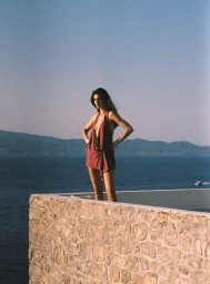 Model Zoi Aggeliki Mantzakanis posing in a mauve halter neck mini dress atop a stone wall overlooking the ocean.