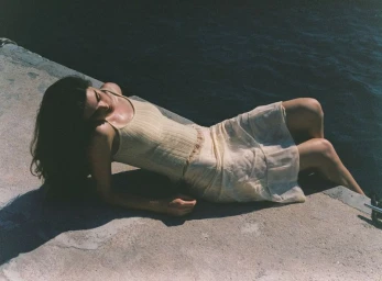 Zoi Aggeliki Mantzakanis reclining on a concrete dock by the ocean wearing a pale yellow knit top and skirt in direct sunlight.