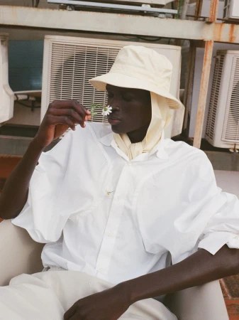 Model Daniel Ajang in a white shirt and cream bucket hat holding a daisy to his nose near air conditioning units.