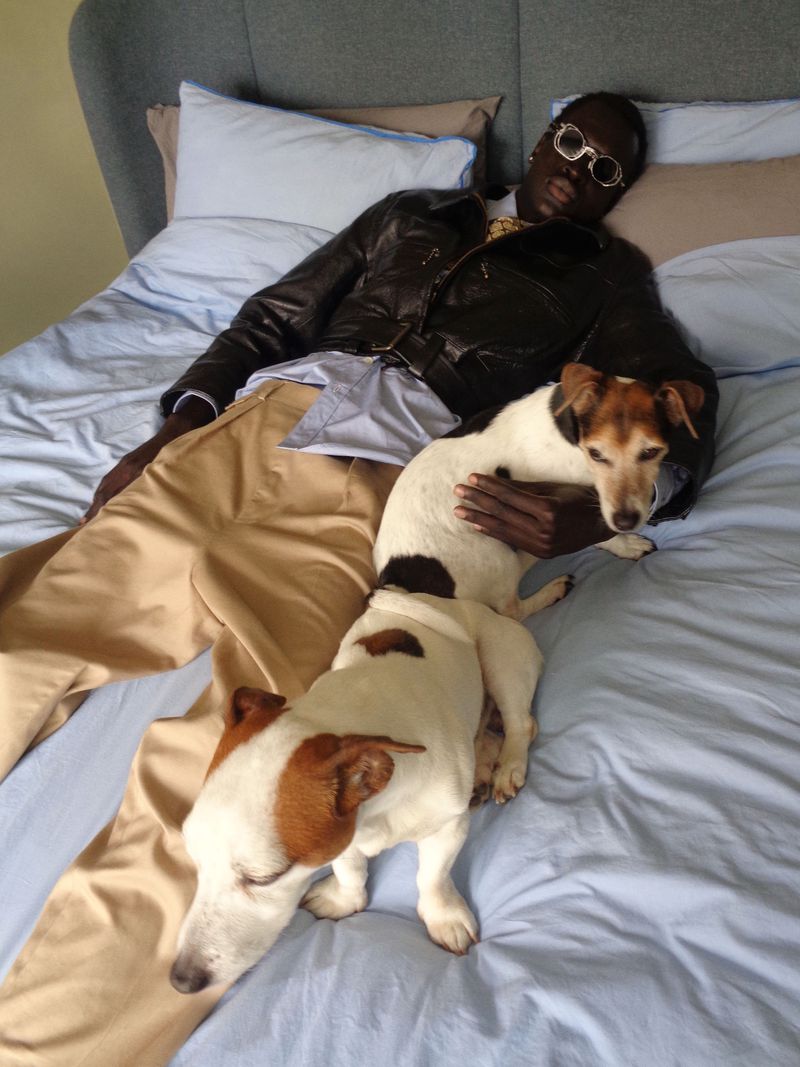 Model Daniel Ajang lies on a bed with two Jack Russell terriers, wearing a dark leather jacket, sunglasses, and tan trousers.