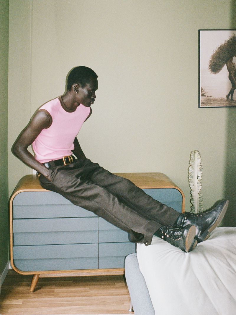 Model Daniel Ajang wearing a pink sleeveless top and brown trousers seated on a mid-century wooden dresser in a bedroom.