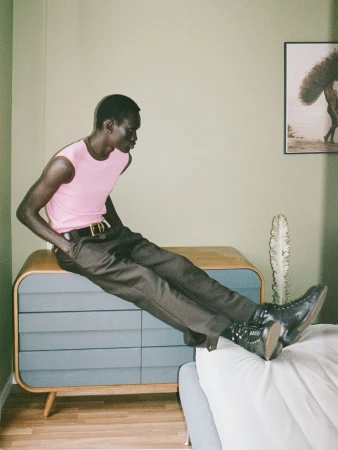Model Daniel Ajang wearing a pink sleeveless top and brown trousers seated on a mid-century wooden dresser in a bedroom.