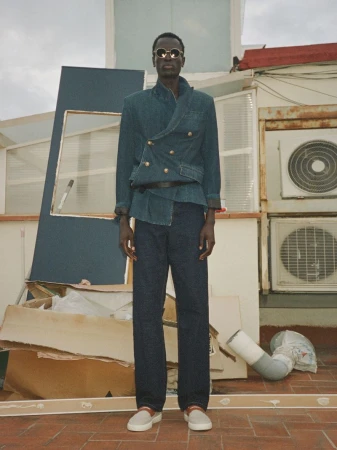Model Daniel Ajang wears a double-breasted denim jacket, dark denim trousers, and sunglasses on an urban rooftop.