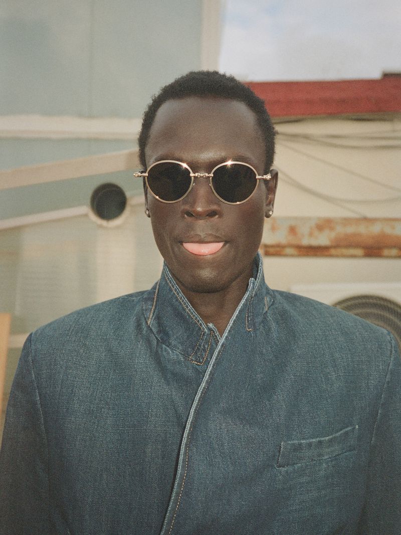 Model Daniel Ajang wearing round sunglasses and a denim jacket while sticking his tongue out outdoors.