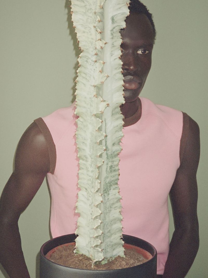 Model Daniel Ajang poses behind a tall potted white cactus wearing a pink sleeveless top against a muted green wall.