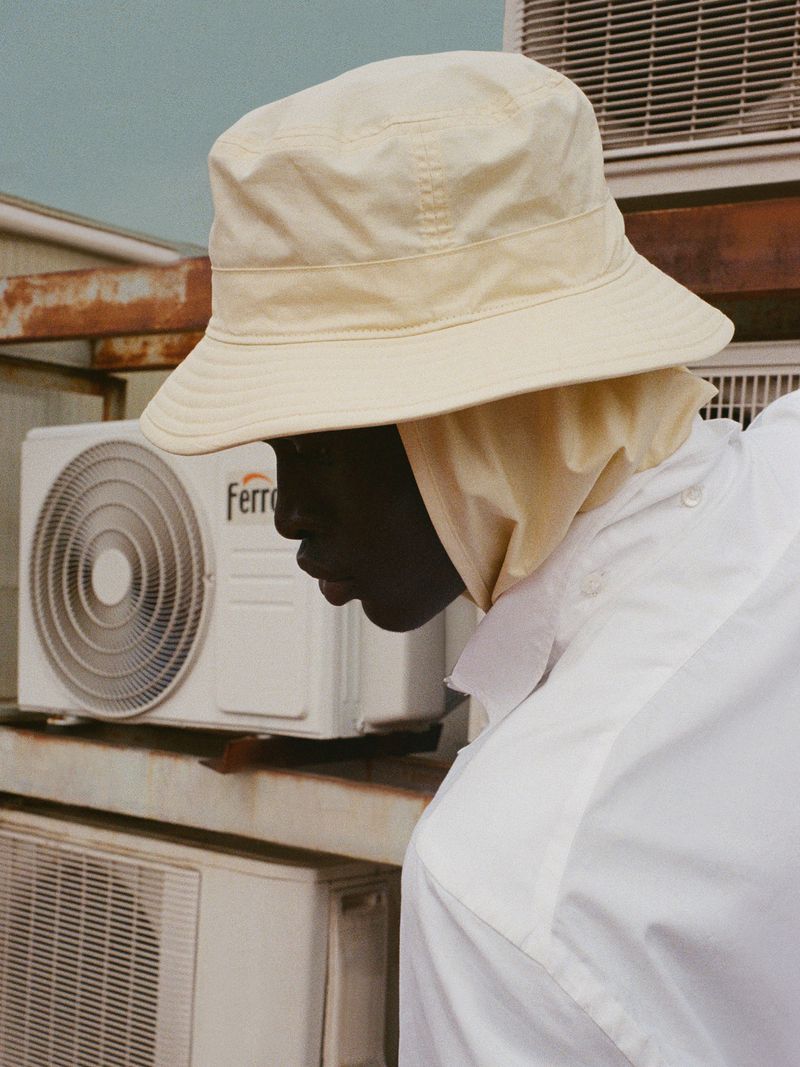 Model Daniel Ajang in profile wearing a cream bucket hat and white collared shirt near air conditioning units.