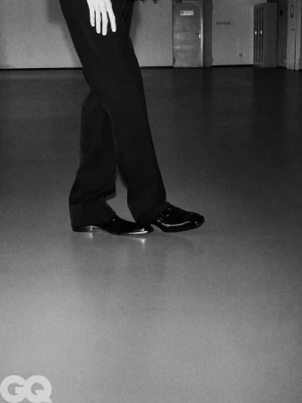 Cropped low-angle view of a model wearing tailored black trousers and polished black dress shoes in a spacious interior.