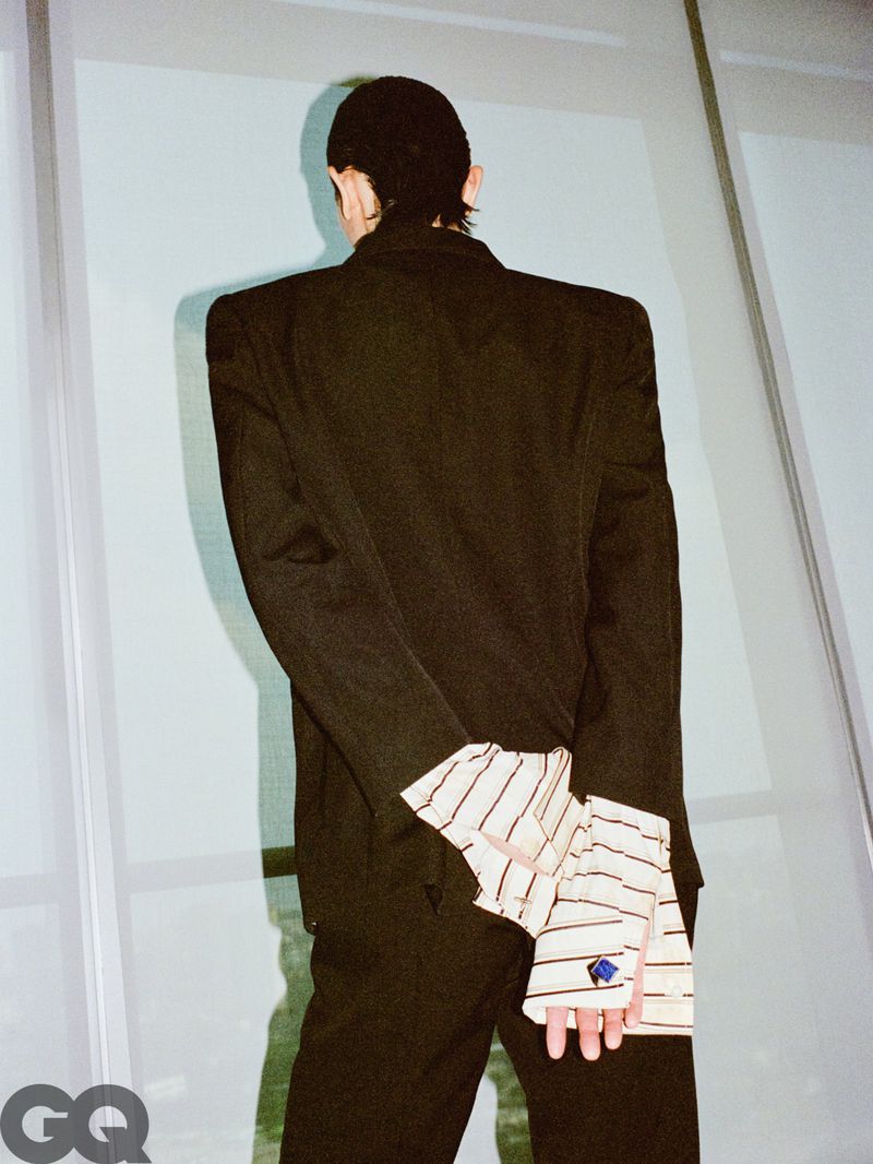 Rear view of a man wearing a structured black blazer with oversized striped cuffs standing against a glass window.