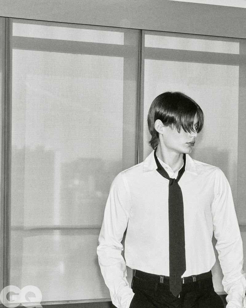 Model Meng Yu Qi in a white collared shirt and loose black tie standing in front of large paneled windows.