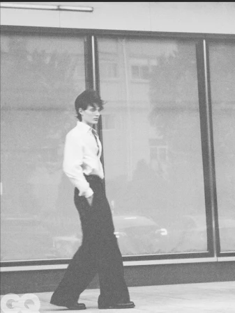 Monochrome editorial photograph of a model walking in a white collared shirt and wide-leg black trousers in front of large glass windows.