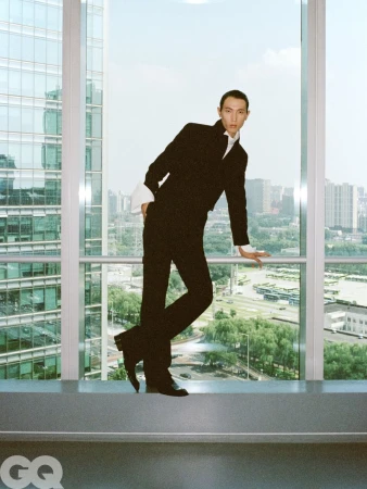 Model Meng Yu Qi wearing a tailored black suit with prominent white cuffs, leaning against a large window overlooking a city skyline.