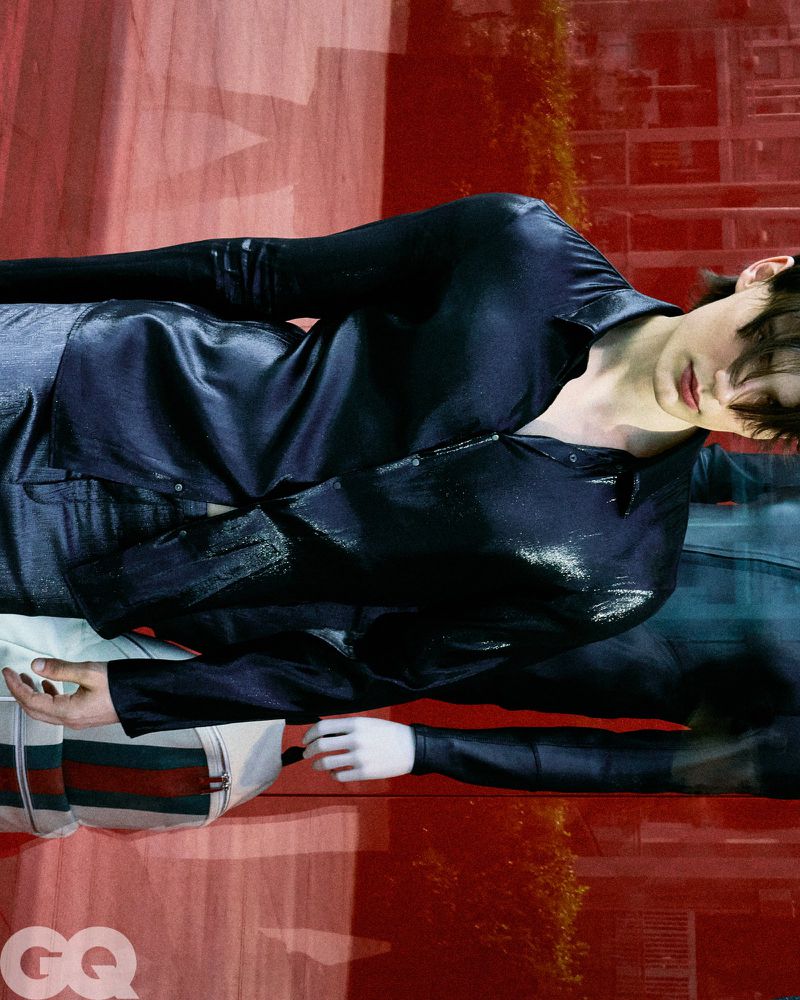 Model Meng Yu Qi wears a dark glossy button-up shirt and carries a striped bag in front of a red-tinted reflective glass window.