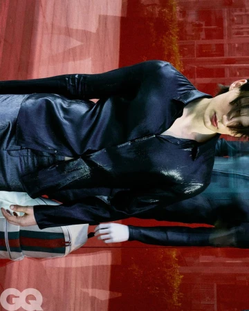 Model Meng Yu Qi wears a dark glossy button-up shirt and carries a striped bag in front of a red-tinted reflective glass window.