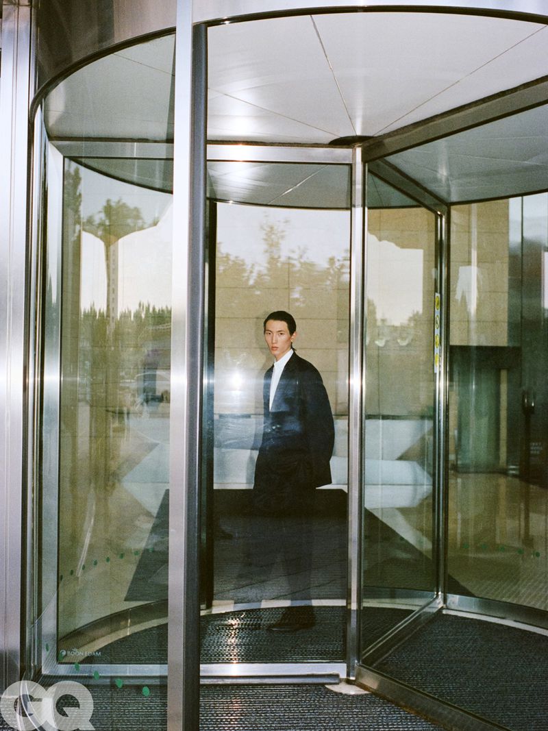 Model Meng Yu Qi wearing a dark suit jacket and white shirt standing inside a glass revolving door entrance.