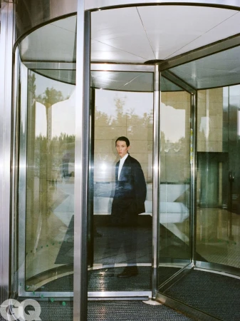 Model Meng Yu Qi wearing a dark suit jacket and white shirt standing inside a glass revolving door entrance.