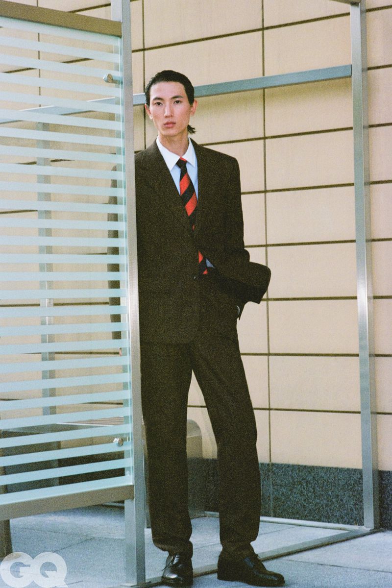 Model Meng Yu Qi wearing a dark tailored suit and striped tie standing beside a glass panel structure.