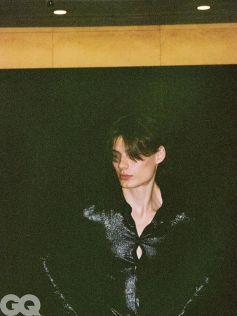Model wearing a shimmering black long-sleeve top looking downward against a dark background in an editorial setting.