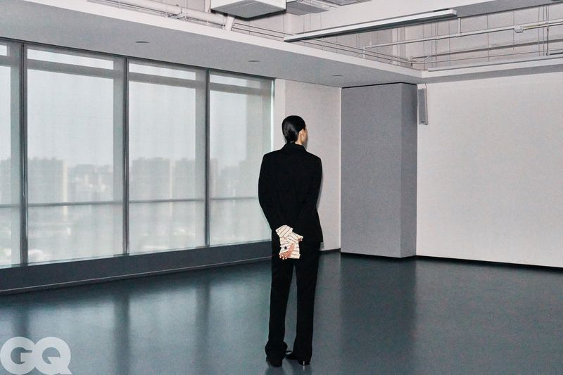 Model Meng Yu Qi seen from behind wearing a black suit with hands clasped behind back in an empty high-rise office space.