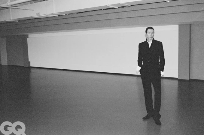Model Meng Yu Qi stands wearing a black tailored suit with white cuffs in a spacious minimalist interior.