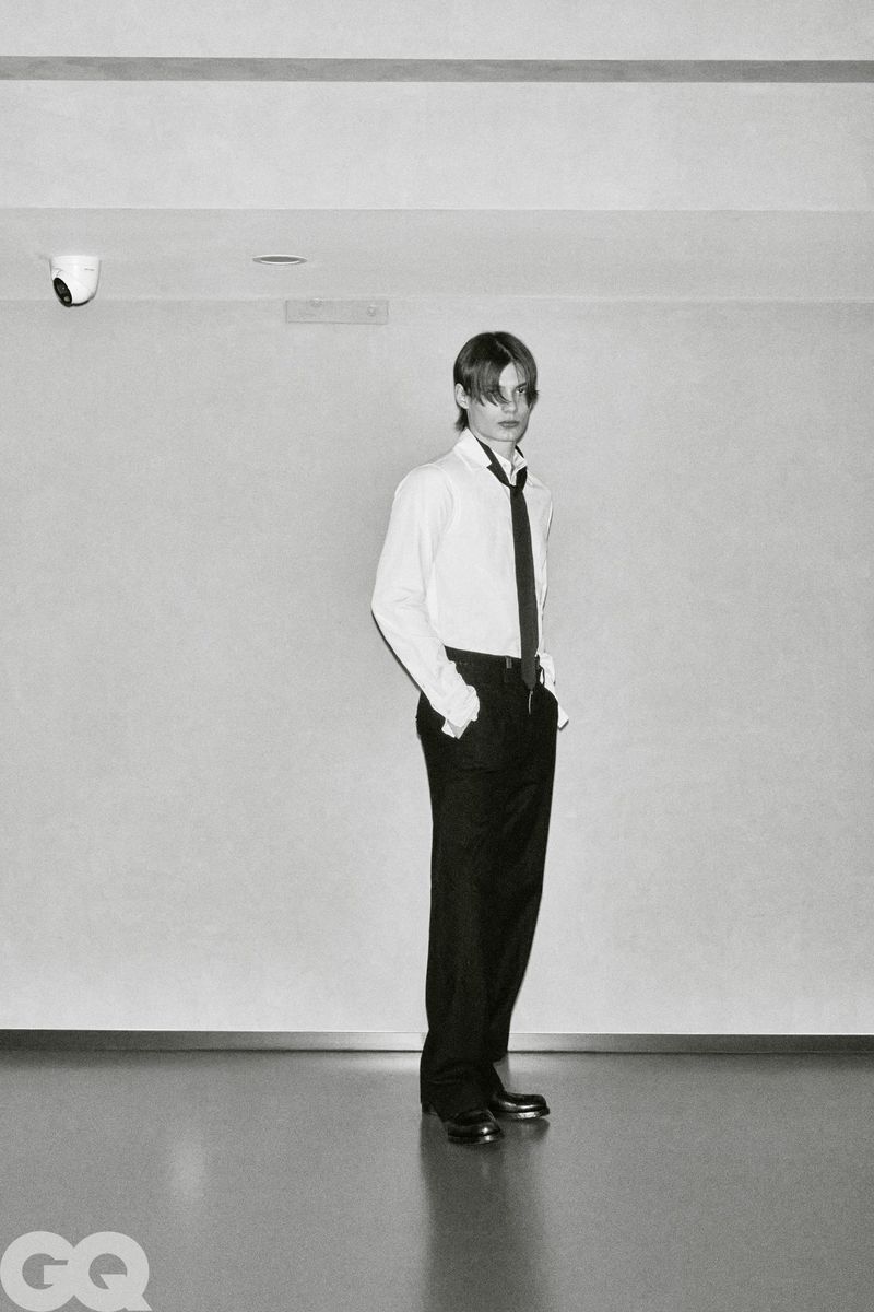 Male model Meng Yu Qi standing in a white collared shirt, black necktie, and dark trousers in a minimalist monochrome setting.